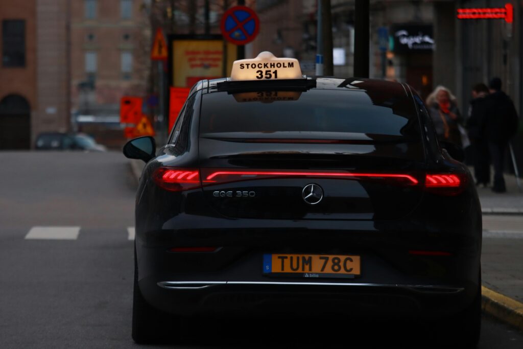 A sleek black Mercedes Benz taxi driving through a lively Stockholm street in the evening glow.
