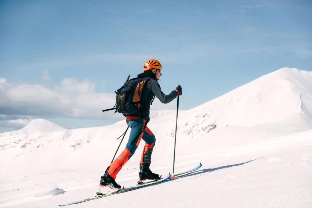 Skier climbing a snowy mountain in bright attire with poles, under a clear blue sky.