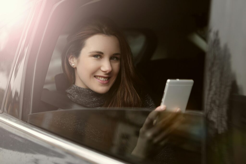 Smiling woman enjoying leisure time in a car using her smartphone on a sunlight day.
